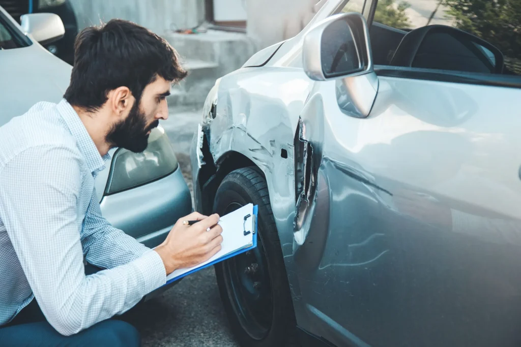 A man holding a clipboard examining damage to a silver car after an accident.