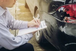 A car after it was rear-ended and someone with a clipboard looking at the back of the car.